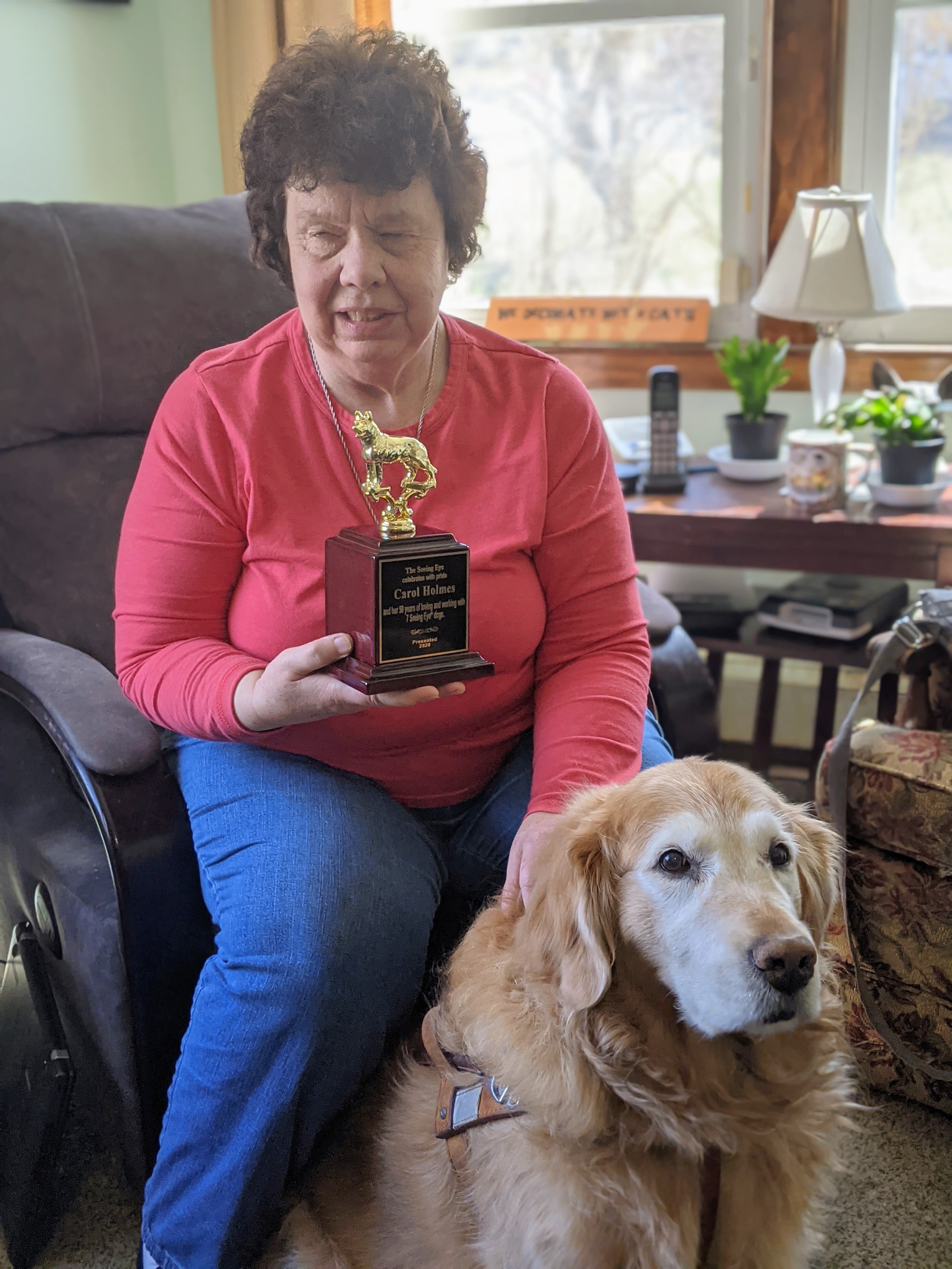 Carol sitting in her living room wearing a red shirt and jeans and sitting next to her is her Golden Retriever Lyric plus she is holding a trophy from The seeing Eye dog on her right hand